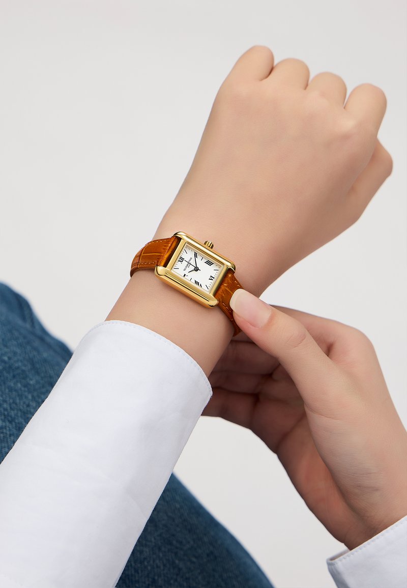 Montre carrée dorée avec un cadran blanc, des chiffres romains noirs et un bracelet en cuir marron, portée sur un poignet à la peau claire.