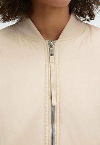 Hellbeige Bomberjacke mit glatter Oberfläche, ausgestattet mit einem silbernen Reißverschluss und einem gerippten Kragen. Keine sichtbaren Muster oder Beschläge.