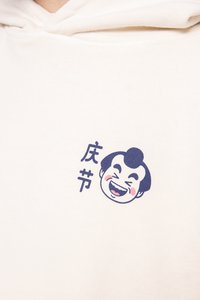 Sudadera crema con un gráfico azul de un personaje sonriente y texto en japonés en el área del pecho izquierdo. Tejido suave con un ajuste relajado.