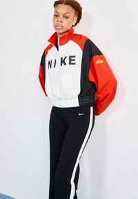 Biela, červená a čierna Nike zipová bunda s výrazným nápisom "NIKE". Skombinovaná s čiernymi nohavicami s bielymi bočnými pruhmi a logom.