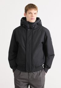Doudoune noire avec une grande capuche, fermeture à glissière à l'avant, boutons-pression et poignets élastiques. Portée par-dessus un pantalon à carreaux gris, elle présente une texture lisse.