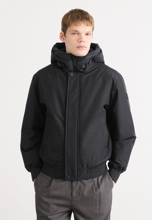 Black puffer jacket with a large hood, zip front, snap buttons, and elastic cuffs. Worn over gray plaid trousers, features a smooth texture.