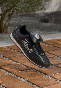 Black sneaker with a glossy finish, featuring a lace-up design, padded tongue, and embossed logo on the tongue, set against a patio background.