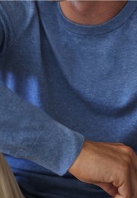 Blå stickad tröja med rund hals och lång ärm, med en mjuk textur och minimala sömmar, mot en beige bakgrund.