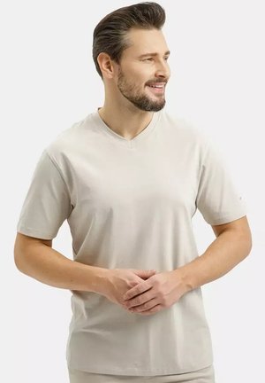 Mężczyzna z krótkimi ciemnymi włosami i brodą, w jasnobeżowej koszulce z dekoltem w serek, stoi ze złożonymi dłońmi i patrzy w prawo.