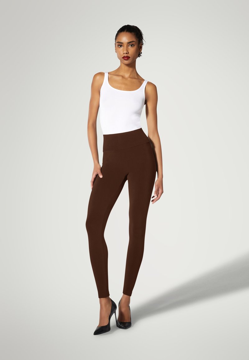 Bruna leggings med hög midja, tillverkade av ett mjukt material, kombinerat med en åtsittande vit linne och svarta högklackade skor.