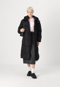 Melnā puffer jaka ar kapuci, ar rāvējslēdzēju un sānu pogām. Valkāta virs rozā grafiskā T-kreklu un melna džinsu svārku. Melnas kurpes un baltas zeķes.