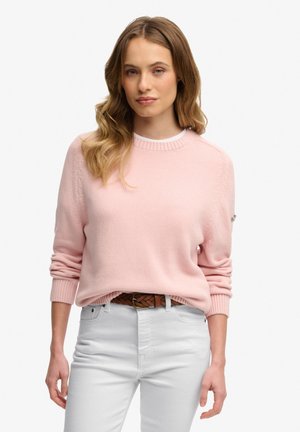 Lys lyserød strikket sweater med rund hals og ribstrikkede ærmekanter, kombineret med hvide jeans og et flettet brunt bælte. Glat tekstur, afslappet design.