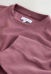 Sudadera burdeos con cuello acanalado, textura suave y detalles de costuras visibles. La etiqueta dice "STUDIO". Presenta una manga doblada.