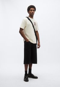 Φωτεινή μπεζ βαμβακερή μπλούζα με γραφικό σχέδιο, συνδυασμένη με χαλαρά μαύρα τζιν σορτς και μαύρα μοκασίνια. Το μοντέλο κρατά μαύρη τσάντα ώμου.