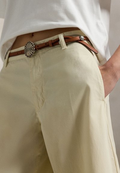 Ανοιχτό κίτρινο βαμβακερό παντελόνι με πλαϊνές τσέπες και εφαρμοστή μέση, συμπληρωμένο με καφέ δερμάτινη ζώνη με ασημί αγκράφα με λουλούδια.