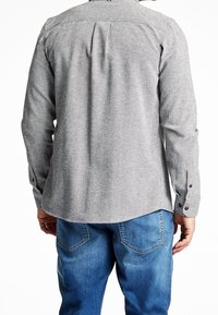 Uomo che indossa una camicia grigia a maniche lunghe con texture e jeans blu, visto di spalle, in piedi contro uno sfondo bianco.