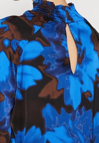 Vêtement à col montant avec un motif floral abstrait bleu et marron, comprenant une petite ouverture en forme de goutte d'eau et une fermeture par un seul bouton à l'arrière.