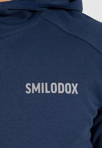 Felpa blu navy con una texture liscia, dotata di un logo grigio "SMILODOX" sul lato sinistro del petto. Design con cappuccio e cuciture a piatto.