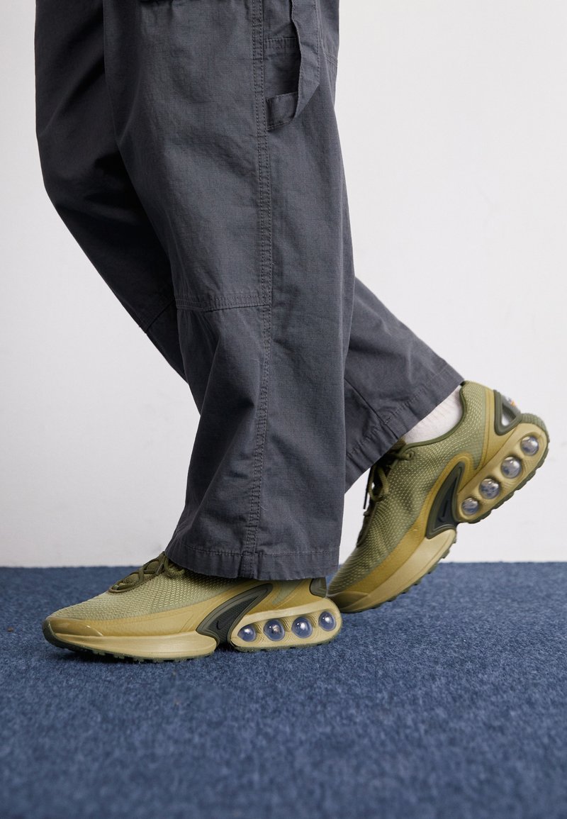 Zapatillas de malla verde con amortiguación redondeada, material texturizado y un diseño aerodinámico. Combinadas con pantalones cargo grises de corte holgado.