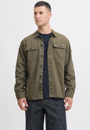 Chaqueta de camisa verde oliva hecha de una mezcla de algodón, que presenta dos bolsillos en el pecho, botones negros y un corte relajado, llevada sobre una camiseta negra.