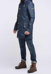 Marinblå regnkappa med en glänsande yta, försedda med huva, guldknappar, sidofickor och en lappar på ärmen. Kombineras med denim och bruna kängor.