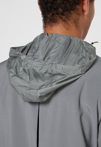 Graue Jacke mit einer strukturierten, leichten Kapuze. Verfügt über einen Rückenlüftungsschlitz für Atmungsaktivität und einen Kordelzug zur Anpassung.