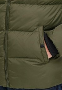Olive green puffer jacket with zipper and snap-button pocket, showing hand partially inserted into pocket over a black ribbed cuff.