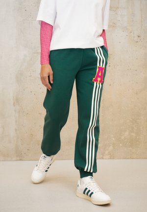 Persona que lleva pantalones deportivos verdes con rayas blancas y parche rosa con la letra A, zapatillas blancas con rayas verdes, camisa blanca y mangas rosas.
