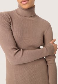 Maglione a collo alto a coste in marrone chiaro, realizzato in tessuto morbido. Presenta maniche lunghe con polsini aderenti e una vestibilità attillata.