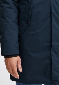 Mørkeblå isolert parkas med en glatt tekstur, som har en sidelomme med en svart knapp og elastiske mansjetter. Synlig hånd på stoffet.