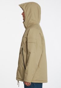 Beige gewatteerde jas met een ritssluiting aan de voorkant, zijzak en textuurstof. Beschikt over een aansluitende capuchon en een losse pasvorm.