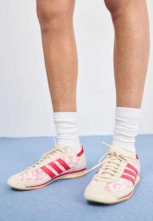 Red and pink patterned athletic shoes with beige accents, featuring three red stripes, laces, and textured soles, worn with white socks.