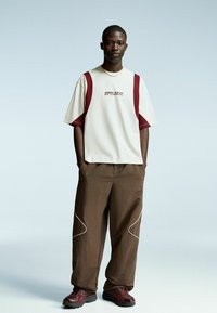 Beige short-sleeve shirt with maroon accents and logo, paired with loose brown pants featuring white stitching details and sleek red sneakers.