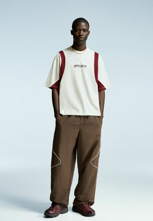 Beige kortærmet skjorte med maroon detaljer og logo, parret med løse brune bukser med hvide syninger og slanke røde sneakers.