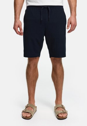 Uomo che indossa pantaloncini neri con coulisse e sandali beige con fibbia, in piedi con le gambe divaricate su sfondo bianco.