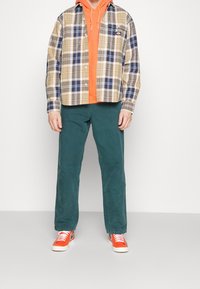 Camicia di flanella a quadri in beige, blu navy e crema sopra un felpa arancione brillante, abbinata a pantaloni in velluto a coste verde e sneakers arancioni.