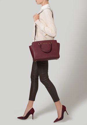 Sac cabas en cuir bordeaux avec ferrures dorées, double poignées supérieures et bandoulière amovible. Le modèle porte un pull crème et un pantalon marron.