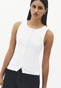 Mujer con una blusa blanca sin mangas con botones y pantalones negros, de pie frente a un fondo liso y claro, mirando hacia adelante.