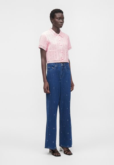 Γυναίκα που φοράει ροζ κοντομάνικο πουκάμισο με κουμπιά και τρύπες, ψηλόμεσο φαρδύ τζιν μπλε με μικρά άσπρα σχέδια και μαύρα σανδάλια με τακούνι.
