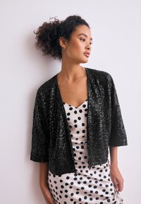 Cardigan noir en sequins avec une coupe ample et des manches mi-longues jusqu'au coude, porté par-dessus une robe crème à pois noirs.