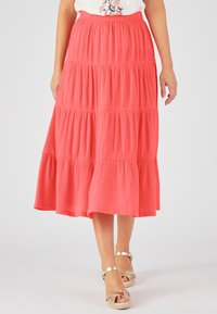 Jupe maxi corail à volants en tissu texturé, taille élastique et silhouette évasée, associée à des sandales métalliques à lanières.
