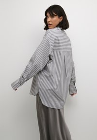 Chemise à boutons rayée gris et blanc avec des manches longues, un pli au dos et un ourlet légèrement arrondi, associée à un pantalon large gris lisse.