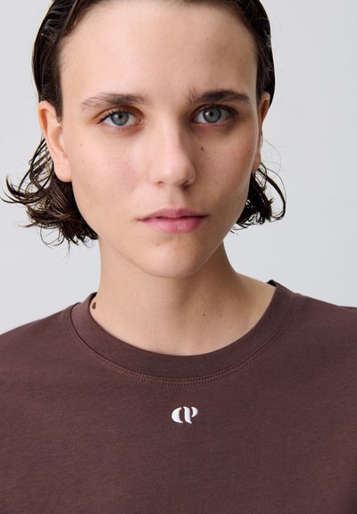 T-shirt en coton marron avec un col rond et un petit logo blanc brodé en haut à gauche. Texture lisse, coupe ajustée.