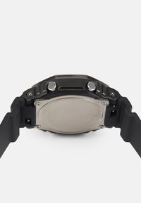 Bracelet de montre en caoutchouc noir avec boîtier métallique, accents argentés, deux boutons sur le côté et un fond circulaire en acier inoxydable.