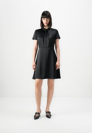 Black short-sleeve dress with a pleated neckline and flared skirt. Smooth texture, fitted bodice, and above-knee length. Black pointed shoes with a strap.