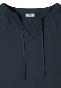 Chemise bleu foncé à col en V avec un cordon de serrage au col et une étiquette blanche portant l’inscription "Cecil" cousue à l’encolure.