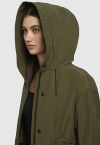 Veste à capuche vert olive avec une capuche matelassée, fermeture éclair noire et boutons-pression. Présente une coupe décontractée et des détails de taille élastiquée.