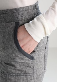 Main dans la poche avant d'un pantalon gris texturé avec ceinture élastique, portant une chemise blanche à manches longues.