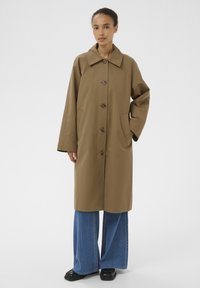 Femme debout portant un long manteau marron boutonné avec un large col, un jean bleu à jambes larges, et des chaussures noires sur un fond clair uni.