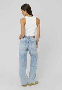 Femme avec de longs cheveux bruns, portant un débardeur blanc, un jean large bleu clair et des baskets vertes, se tenant de dos avec les mains dans les poches.