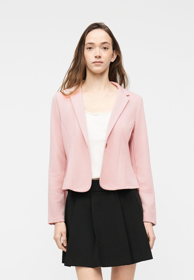 Jeune femme portant un blazer rose clair sur un haut blanc, associé à une jupe plissée noire, debout devant un fond uni.