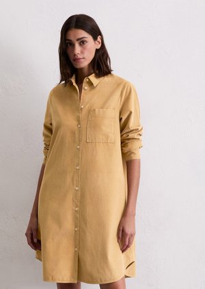 Vrouw met schouderlang donker haar draagt een losse, langemanige beige overhemdjurk met een borstzakje tegen een witte muur.