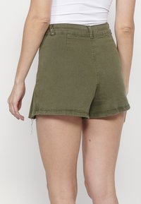 Femme portant un short taille haute vert olive avec un ourlet brut, vue de dos, et un haut blanc sans manches.