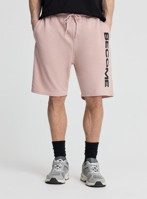 Uomo che indossa pantaloncini rosa con la scritta "BECOME" sulla gamba destra, calzini neri, sneaker grigie e una maglietta nera, mani nelle tasche.